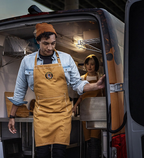 Uniformed worker standing in back of van with doors open and tools and parts around him.