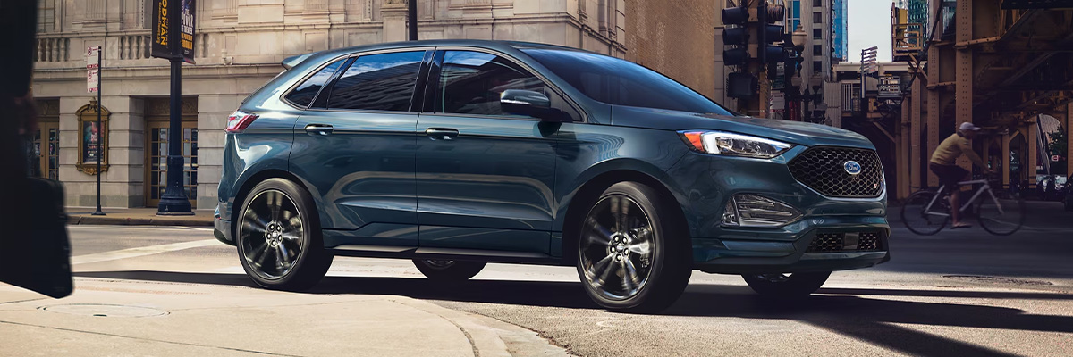 A 2024 Ford Edge® SUV in Stone Blue being driven around the curve in a city street
